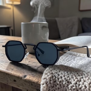 May include: Black octagonal sunglasses with dark lenses rest on a wooden table. A white mug with steam rising from it sits in the background, along with an open book and a knitted blanket. The sunglasses have a sleek, modern design.