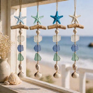 Peut inclure: Quatre décorations côtières suspendues. Chacune présente une étoile de mer dans des tons de bleu et de blanc, fixée à une branche de style bois flotté. En dessous, des morceaux de verre bleus, verts et blancs, avec un coquillage en bas. L'arrière-plan montre une plage et l'océan.