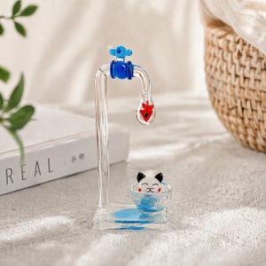 May include: A clear glass water faucet sculpture with blue and red accents. The faucet has a blue handle and a red teardrop-shaped element. A small bowl with a cat face is at the base, with blue liquid inside.