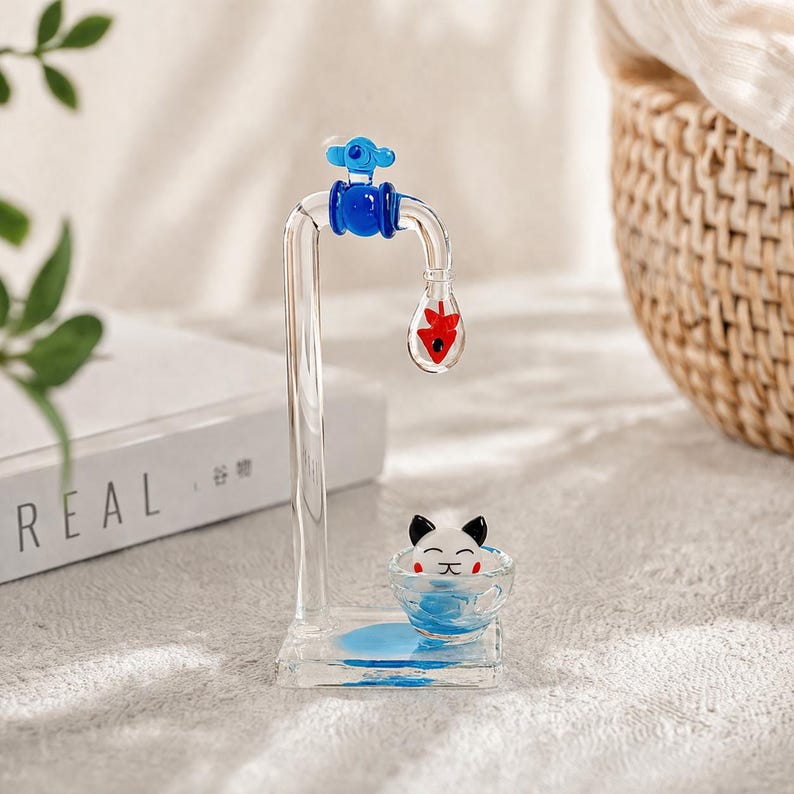 May include: A clear glass water faucet sculpture with blue and red accents. The faucet has a blue handle and a red teardrop-shaped element. A small bowl with a cat face is at the base, with blue liquid inside.