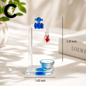May include: A clear glass faucet sculpture with blue and red accents. The faucet is 3.35 inches tall and 1.42 inches wide. The faucet has a blue handle and a red teardrop-shaped element. A small glass bowl sits below the faucet.
