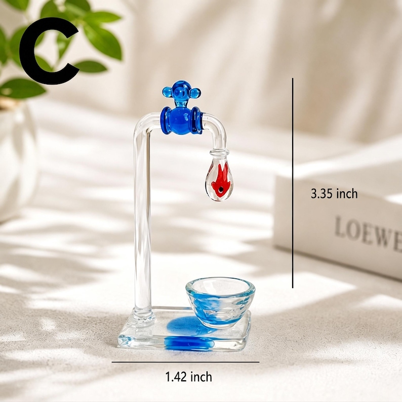 May include: A clear glass faucet sculpture with blue and red accents. The faucet is 3.35 inches tall and 1.42 inches wide. The faucet has a blue handle and a red teardrop-shaped element. A small glass bowl sits below the faucet.