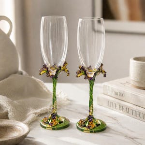 Puede incluir: Dos copas de champán con cuencos de cristal transparente y tallos verdes. Los tallos están decorados con diseños florales morados y amarillos. Las copas están sobre una superficie blanca.