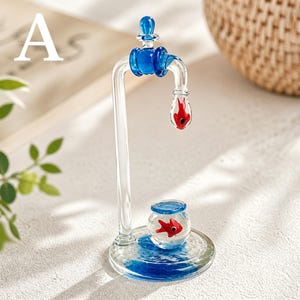 May include: A clear glass sculpture of a faucet with blue accents and a red fish inside a water droplet. A small fishbowl with a red fish sits below the faucet, on a blue water-like base. The sculpture is a decorative item.