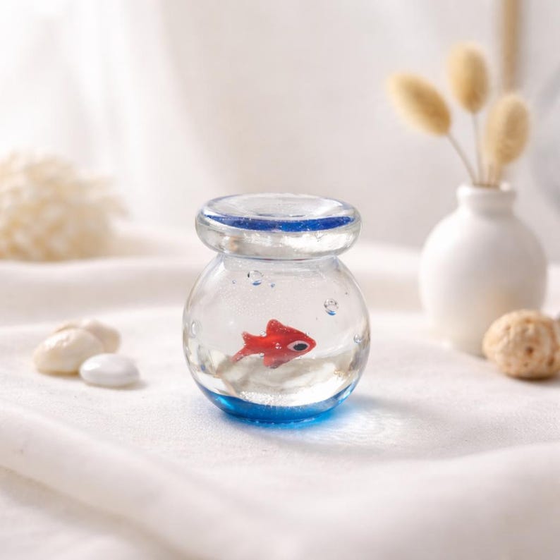 May include: A small, clear glass jar with a blue rim and base, containing a red fish figurine. The jar has a lid and sits on a white surface. Decorative elements include a small white vase with dried flowers and a few white stones.