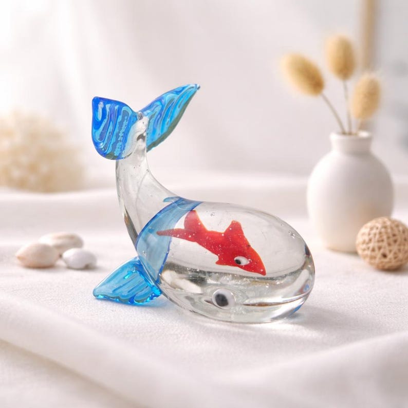 May include: A clear glass whale figurine with blue accents on the fins and tail. Inside the whale is a red fish. The figurine is on a white surface.
