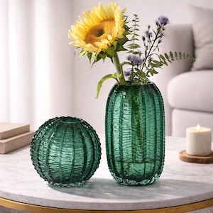 Peut inclure: Deux vases en verre vert, un rond et un grand, avec un design texturé et nervuré. Le grand vase contient un tournesol et d'autres fleurs. Une bougie allumée est posée sur un dessous de verre en bois à proximité. Les vases sont sur une table en marbre blanc.