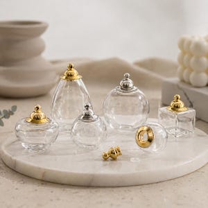 May include: A collection of clear glass perfume bottles with gold and silver-colored lids. The bottles are various shapes, including round, teardrop, and square. The bottles are arranged on a white marble tray, with a few loose gold-colored stoppers.