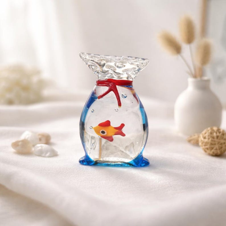 May include: A clear glass fish-shaped ornament with a red starfish and a yellow and orange fish inside. The ornament has a blue base and a red ribbon around the neck. Decorative accents include a small white vase and a woven ball.
