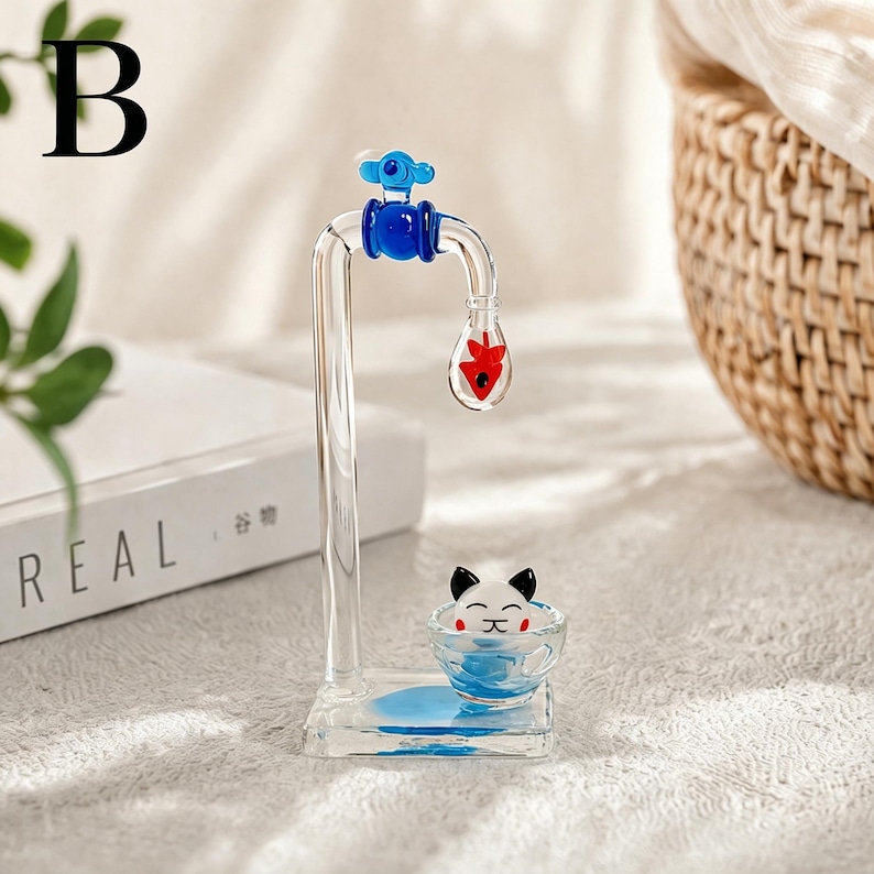 May include: A clear glass sculpture of a faucet with blue and red accents. A small bowl with a cat face is at the base. The sculpture has the letter "B" in the upper left corner.