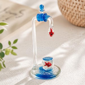 May include: A clear glass sculpture of a faucet with blue accents. A red fish is suspended in the faucet's spout, and another red fish swims in a small glass bowl below. The base has blue water-like details.