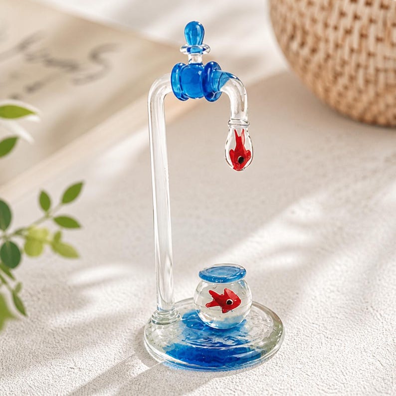 May include: A clear glass sculpture of a faucet with blue accents. A red fish is suspended in the faucet's spout, and another red fish swims in a small glass bowl below. The base has blue water-like details.