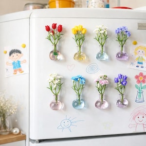 May include: A collection of small, glass bud vases with colorful flowers attached to a white refrigerator door. The flowers include red tulips, yellow tulips, white, blue, pink, and purple blooms. Children's drawings are also attached to the refrigerator.
