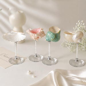 Pode incluir: Quatro cálices decorativos de conchas com hastes e bases transparentes. As conchas são brancas, rosa e verdes, e estão dispostas sobre uma superfície branca. Um pequeno cartão e flores brancas estão no fundo.