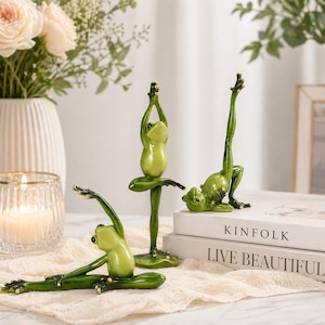 May include: Three green frog figurines in yoga poses. The frogs have gold accents and are positioned on a white surface. One frog is in a standing pose, another is in a seated pose, and the third is in a backbend. The figurines are decorative.