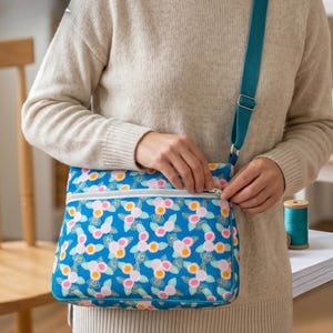 May include: A rectangular crossbody bag featuring a vibrant blue background with a repeating pattern of pink and orange circular floral motifs and green leaves. The bag includes a white zippered front pocket and a solid teal adjustable shoulder strap.