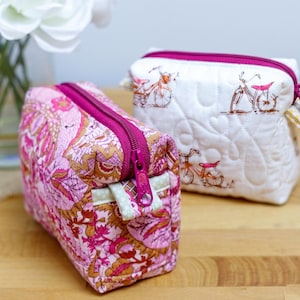 May include: Two small, rectangular cosmetic bags. One is patterned with pink and gold floral designs, and the other is white with a bicycle print. Both bags have a bright pink zipper closure.