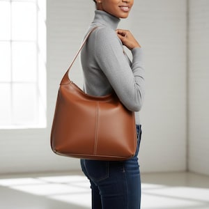 May include: A brown leather shoulder bag with a curved shape and a long strap. The bag has a vertical seam down the center and a zipper closure. The bag is being carried by a person wearing a gray turtleneck and blue jeans.