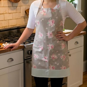 May include: A kitchen apron featuring a brown and white gingham check pattern overlaid with a repeating floral print of pink roses. The apron includes a solid light green and white striped hem at the bottom and matching neck and waist ties.