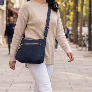 Puede incluir: Un bolso bandolera de mezclilla azul oscuro con forma rectangular, un bolsillo frontal con cremallera y tirador de cuero marrón, y una correa ajustable a juego. El bolso se lleva a la altura de la cadera con herrajes plateados.