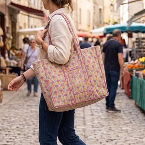 Può includere: Una grande borsa tote trapuntata rettangolare con un motivo floreale ripetuto nei toni del rosa, giallo e verde su sfondo chiaro. La borsa è dotata di due tracolle e di una tasca rettangolare verticale sul pannello frontale esterno.
