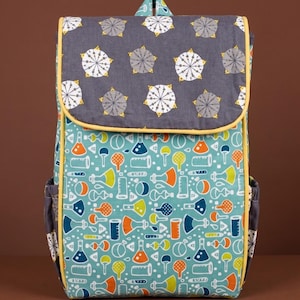 Puede incluir: Una mochila colorida con cierre de solapa. La solapa es gris con un patrón de estrellas blancas y amarillas. El cuerpo principal es turquesa con un patrón repetido de equipos de laboratorio en naranja, amarillo y azul. La mochila tiene ribetes amarillos y bolsillos laterales.