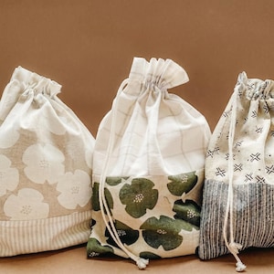 May include: Three drawstring bags with various fabric patterns. One bag features large white floral designs on a beige background, another has green floral patterns, and the third has a black and white geometric pattern. Each bag has a neutral-colored drawstring.