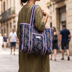 May include: A dark blue tote bag featuring a gold paisley pattern, accented with light purple piping along the edges and straps. The bag includes a rectangular white label with black text on the front pocket and two long shoulder straps.