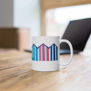 May include: A white ceramic mug with a black handle and the text "But First, Coffee" in a bold, black font. The mug is set against a blurred background. The mug appears to be approximately 10 cm tall.