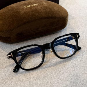 May include: Black Tom Ford spectacles featuring a rectangular frame and silver-tone T-shaped detailing on the temples. The glasses are presented with a brown velvet-textured hard case, which features the Tom Ford logo embossed in gold on the lid.