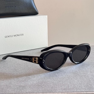 May include: Black oval-shaped sunglasses featuring dark lenses and a gold-toned metal logo detail on the temple. The frame includes three small silver-toned dots on the outer corners. The brand name Gentle Monster is printed on the temple and box.