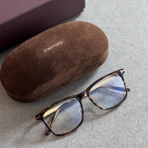 May include: Tortoiseshell-patterned eyeglasses with a brown suede case and a dark purple box, all branded with "TOM FORD". The glasses feature a rectangular frame design.