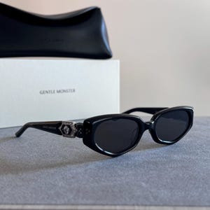 May include: Black sunglasses with dark lenses and a thick frame. The temples feature a silver-toned hexagonal detail embedded with small clear crystals and the text GENTLE MONSTER. A white box with the text GENTLE MONSTER and a black case are behind.