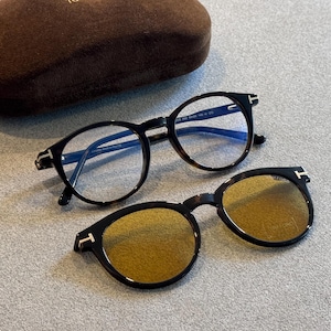 May include: Two pairs of eyeglasses with a tortoiseshell pattern and black frames. One pair has clear lenses, and the other has yellow-tinted lenses. A brown glasses case is in the background.