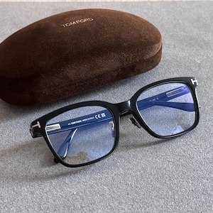 May include: Black Tom Ford spectacles with rectangular frames and blue-tinted lenses, featuring a silver T-shaped logo on the temples. The glasses are accompanied by a brown velvet-textured hard case with the Tom Ford logo embossed on the top.