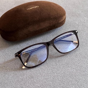 May include: Dark brown tortoiseshell spectacles with a rectangular frame and a gold-tone T-shaped hinge detail. The glasses feature clear lenses and are accompanied by a brown velvet hard-shell case with the gold-embossed text TOM FORD.