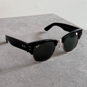 May include: Black sunglasses with a gold-colored lower frame and dark lenses. The sunglasses have a classic design with a thick, black frame and the Ray-Ban logo in white on the temple.