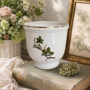 May include: A white ceramic planter with a flared rim and a pedestal base. The exterior features a botanical illustration of a green leafy branch with small brown tendrils. The rim and the base are both accented with a thin, metallic gold trim.