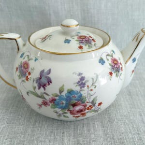 May include: A white porcelain teapot featuring a colorful floral pattern with pink, blue, and purple blossoms. The teapot has a matching lid with a small knob, a curved handle, and a spout, all accented with thin gold trim along the edges.