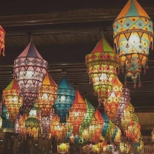 May include: A collection of hanging fabric lanterns in various colors including pink, orange, blue, green, and yellow. Each lantern features a tiered, geometric design with intricate stitched patterns and small decorative tassels.