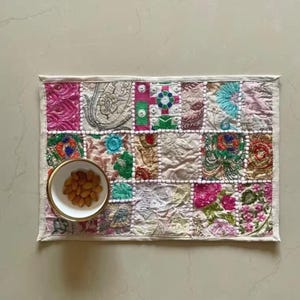May include: A rectangular placemat featuring a patchwork of embroidered fabric squares in various colors including pink, green, blue, and cream, with white pom-pom trim between sections. A small white bowl filled with brown almonds sits on the left.