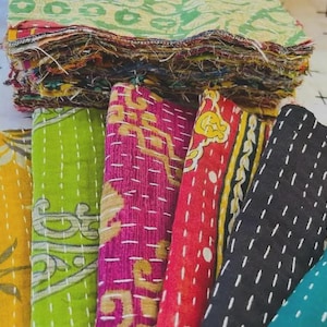 May include: A stack of rectangular fabric swatches featuring various colors including yellow, lime green, magenta, red, black, and teal. Each piece of fabric displays a distinct pattern and is held together by visible white running stitch embroidery.