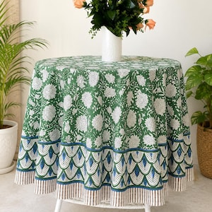 Könnte beinhalten: Eine runde Tischdecke mit grünem Hintergrund und weißem Blumenmuster. Der untere Rand zeigt ein sich wiederholendes fächerförmiges Motiv in Blau, Grün und Weiß, abgeschlossen mit einem schmalen braun-weiß gestreiften Saum am unteren Ende.