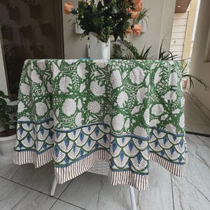 Pode incluir: Uma toalha de mesa redonda com fundo verde e um padrão floral branco estampado. A borda apresenta um design festonado em azul e verde sobre uma bainha fina com listras marrons e brancas, cobrindo uma pequena mesa branca.