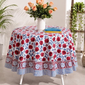 Può includere: Una tovaglia rotonda con sfondo bianco e un motivo ripetuto di fiori rossi e foglie blu. Il bordo presenta una fascia floreale rossa seguita da un orlo a righe blu e bianche. Una brocca bianca con rose color pesca è posizionata sopra.
