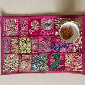 May include: A rectangular placemat featuring a patchwork of vibrant pink fabric panels with intricate gold, green, and blue embroidery, floral patterns, and metallic beadwork. A small white bowl containing brown almonds sits on the top right corner.