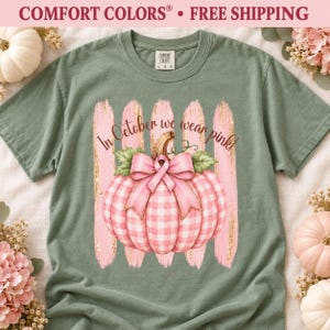 Op de afbeelding: Een saliegroen T-shirt met korte mouwen en een afbeelding van een roze-witte pompoen met een roze strik. De tekst "In October we wear pink!" staat boven de pompoen gedrukt over vijf verticale penseelstreken in een roze kleur.