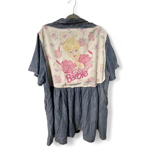 Peut inclure: Une chemise en jean bleu avec un grand panneau arrière couleur crème orné d'une impression graphique représentant un personnage blond et le texte Barbie. Le motif inclut des roses roses, des cœurs et des notes de musique. Manches courtes.