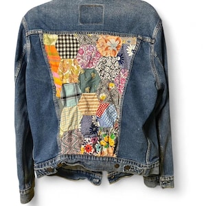 Puede incluir: Chaqueta vaquera azul con un diseño de patchwork en la espalda. El patchwork presenta varios patrones florales y geométricos en tonos naranja, rosa, verde y azul. La chaqueta tiene un cuello clásico y cierres de botón.