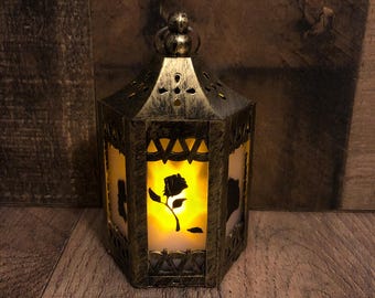 May include: A hexagonal lantern with a distressed gold finish and a metal frame featuring cut-out patterns. The lantern has translucent panels displaying a black silhouette of a rose and emits a warm, glowing yellow light from within.